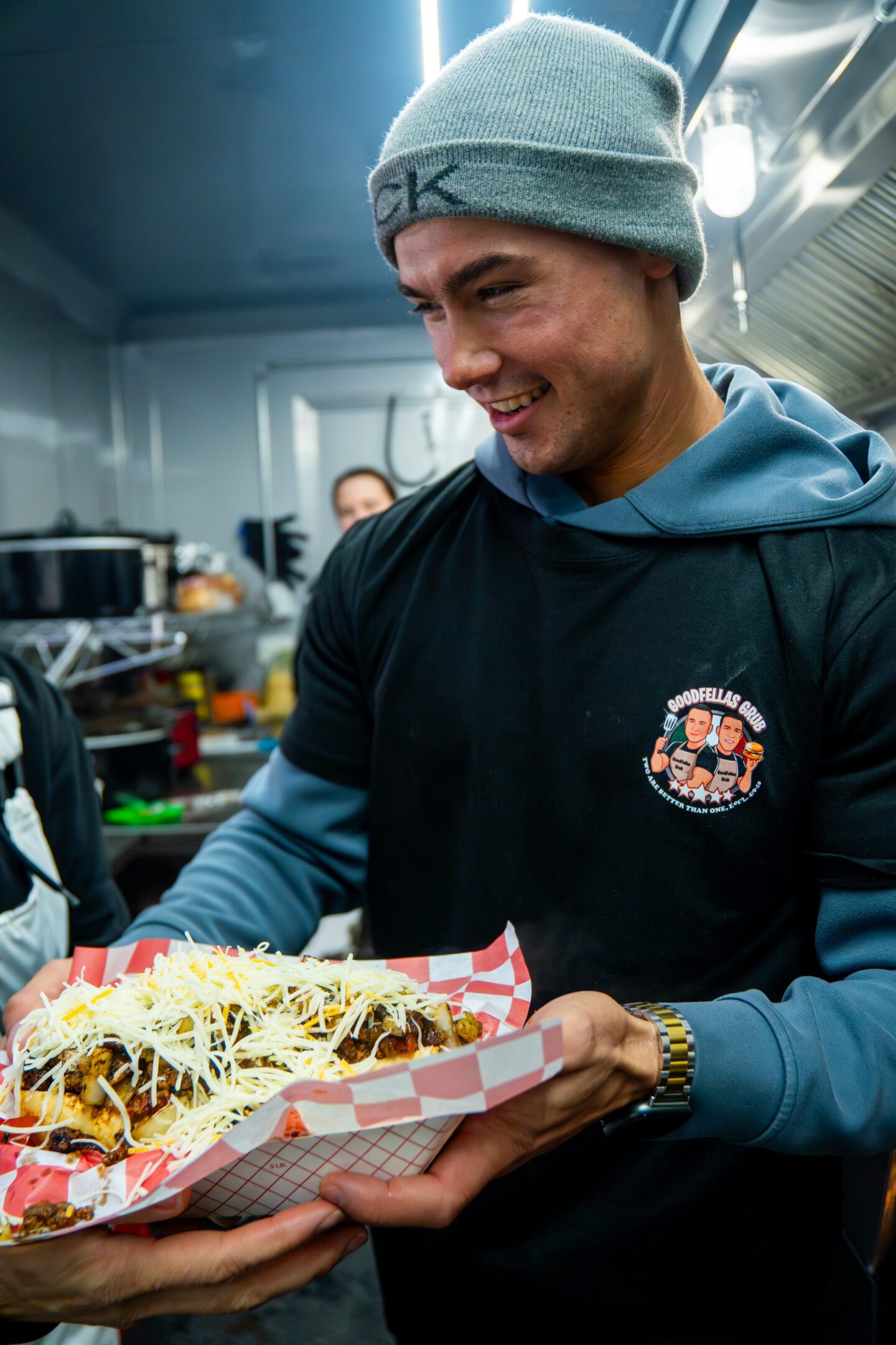 Smiling man holding a loaded sandwich tray in a casual setting.