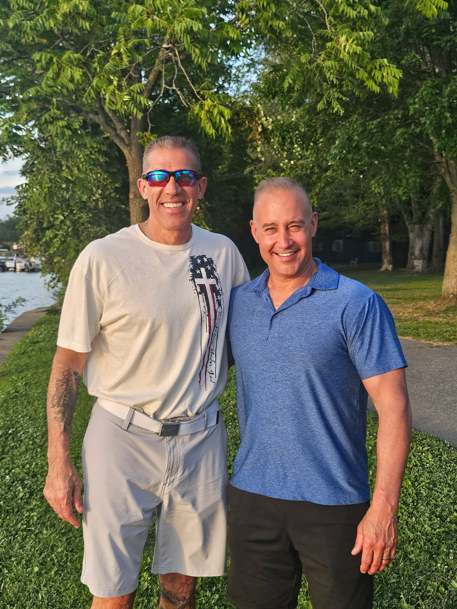 Two men smiling outdoors with trees and water in the background.